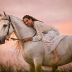 A serene photo of a woman with long dark hair, adorned in a flowing white dress, gracefully sitting bareback on a pristine white horse in a sun-drenched field.