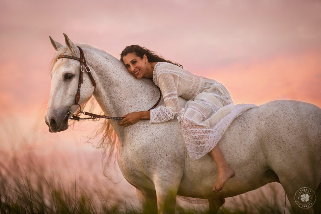 A serene photo of a woman with long dark hair, adorned in a flowing white dress, gracefully sitting bareback on a pristine white horse in a sun-drenched field.