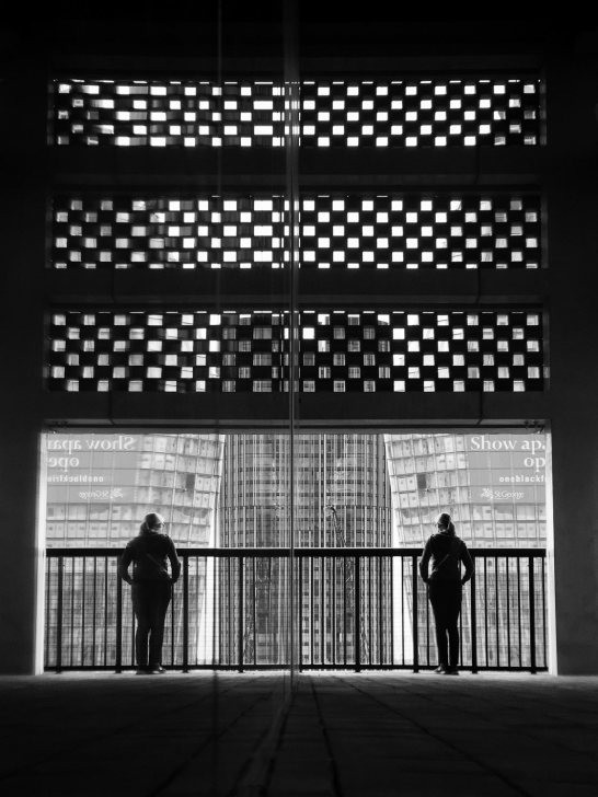 Black and white photograph of a person dwarfed by a large building, suggesting a story of scale and isolation