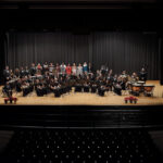 alt text: A photo of an orchestra on a stage with a conductor in the center.