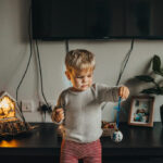 A young boy is holding red Christmas baubles, standing in front of a Christmas tree, bathed in natural light from a nearby window.