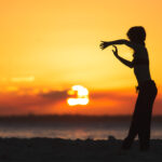 alt text: A dancer poses on a beach with a large sun in the background.