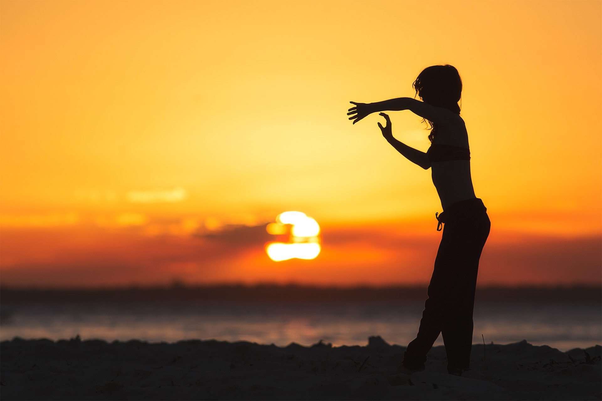 alt text: A dancer poses on a beach with a large sun in the background.