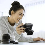 woman in white swaeter holding a camera and looking at her laptop