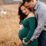 expecting parents standing in a field of golden grasses as dad hugs mom from behind during a maternity photoshoot