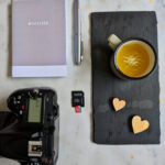 Overhead still life photography shot of a camera, notebook, pen, coffee cup and memory card