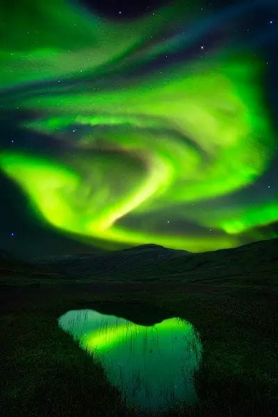 Vibrant aurora borealis display over Icelandic landscape, showcasing strong green and red colors with reflections in water.