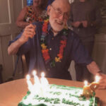 Scan of a joyful nonagenarian celebrating his 94th birthday with family. He is holding up a hand in cheer, wearing a flower lei, and smiling broadly at a birthday cake marked '94'. Two younger family members flank him, sharing in the joyous occasion in a cozy living room setting.