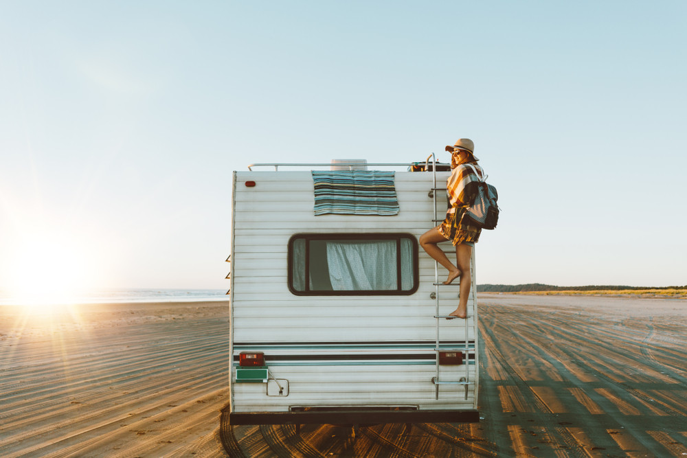 Smiling woman in hat standing near a vintage RV on a sunny day