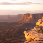 Elevated perspective of a blonde woman in a black jacket and blue jeans, sitting on a high precipice in Canyonlands National Park, Utah, USA, capturing a daring moment with a safe setup