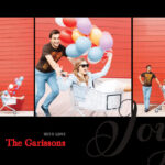 A couple playfully poses with a shopping cart and colorful balloons in front of a red wall.