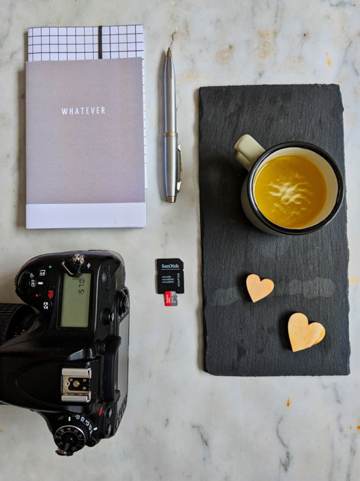 Overhead view of camera, notebook, pen, coffee cup, and SD card, illustrating the photographer's tools.