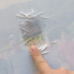 Hand gently rubbing wet paper fibers off the wood surface to reveal the transferred photo, showcasing the delicate process of decoupage