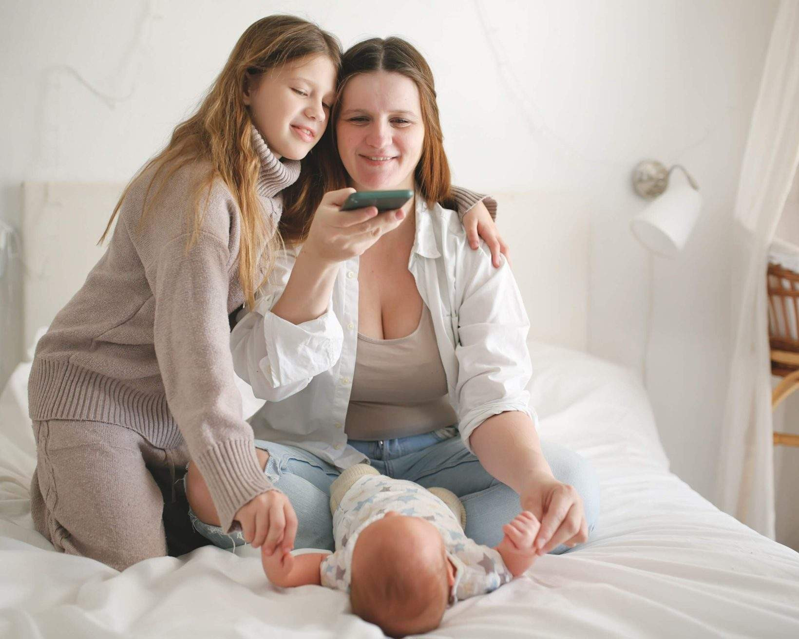 Mother and older daughter take pictures on phone of baby on bed in cozy real bedroom with wicker cradle