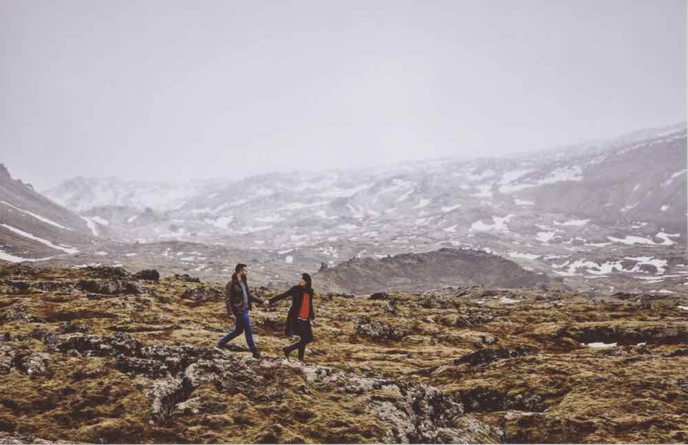 Scan of a photo: Beautiful icelandic landscape featuring a couple walking towards the right, centered in the photo. She’s leading him and they’re holding hands and she looks back and gazes into his eyes. She’s wearing black pants, leather boots, and a long navy sweater with a red shirt under it. He’s wearing a leather jacket, grey shirt, blue pants, and casual green leather sneakers. There’s stark contrast between the rocky and green mossy area they’re walking on and the hill behind them which has a white, almost-snowy overlay. The hillside is half covered in snow.