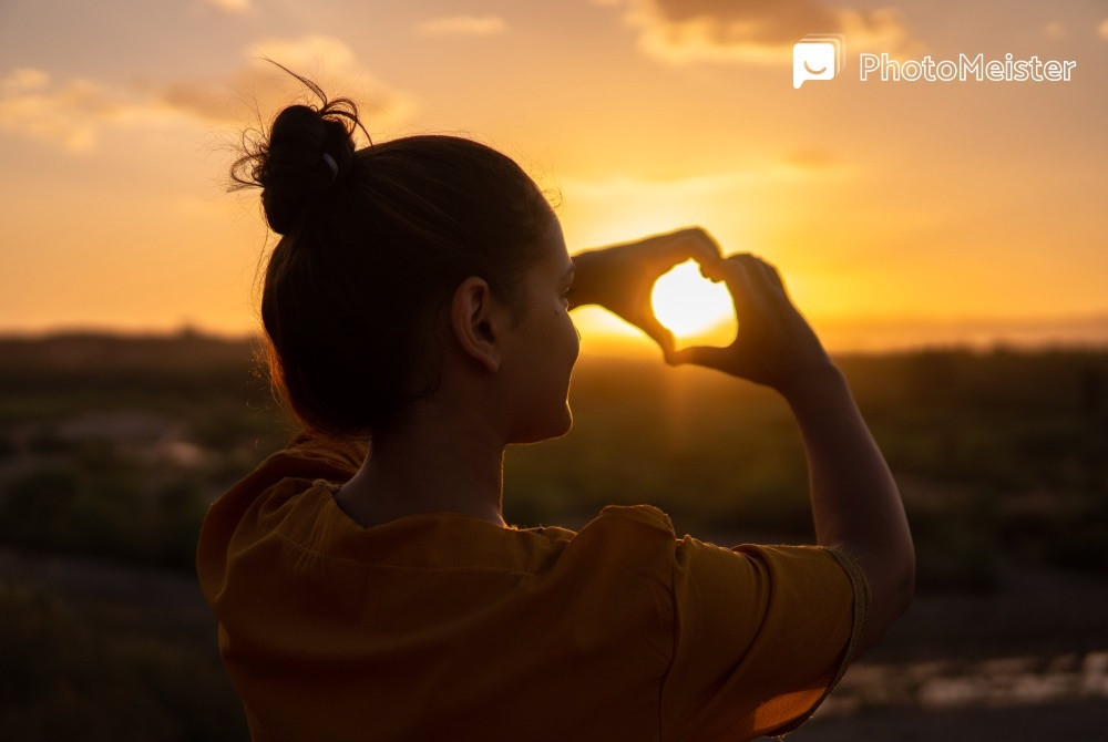 A person uses a smartphone with a solar filter attached to the camera lens to photograph the sunset, while wearing solar eclipse glasses for eye protection.